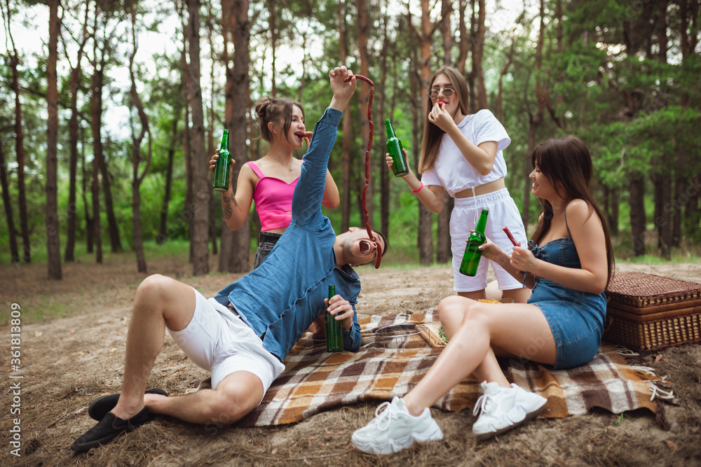 Youth. Group of friends clinking beer bottles during picnic in summer forest. Lifestyle, friendship, having fun, weekend and resting concept. Looks cheerful, happy, celebrating, festive.
