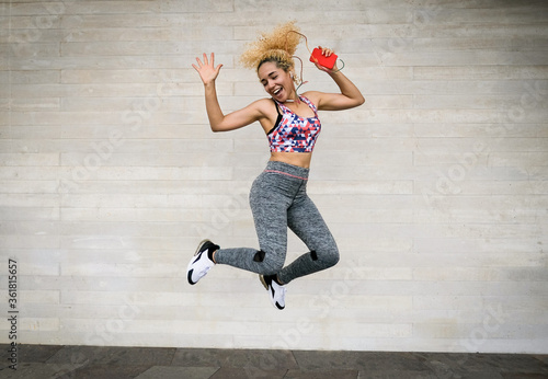 Young multiracial woman listening music from playlist and jumping - Happy sportive woman dancing with headphones and holding smartphone in her hands