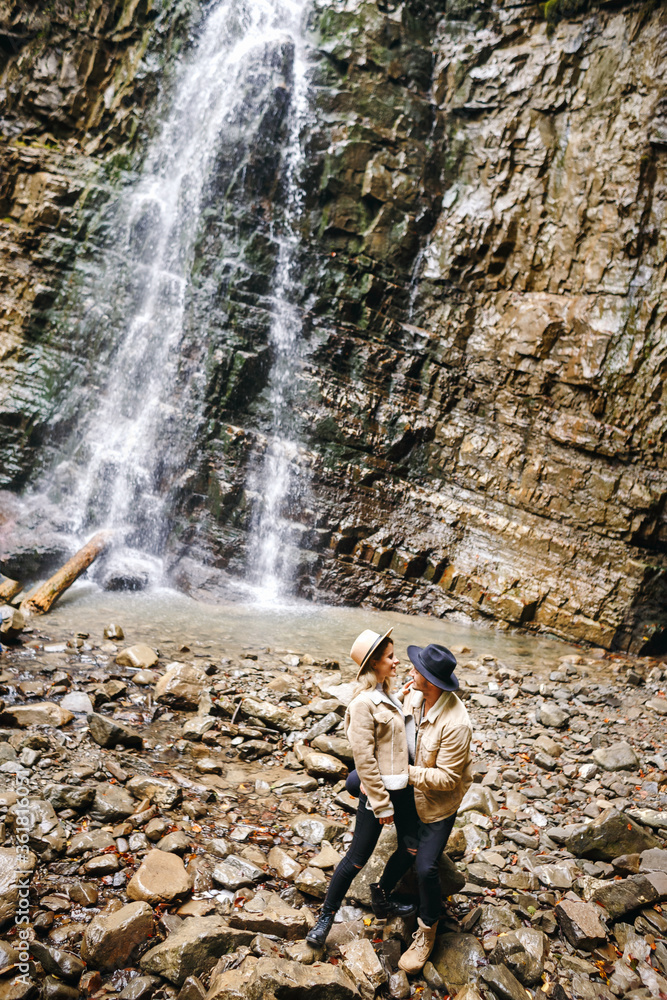 Obraz premium Young and beautiful couple at the mountain waterfall - Happy tourists visiting mountains. Lovestory. Tourists in hats. Military fashion.