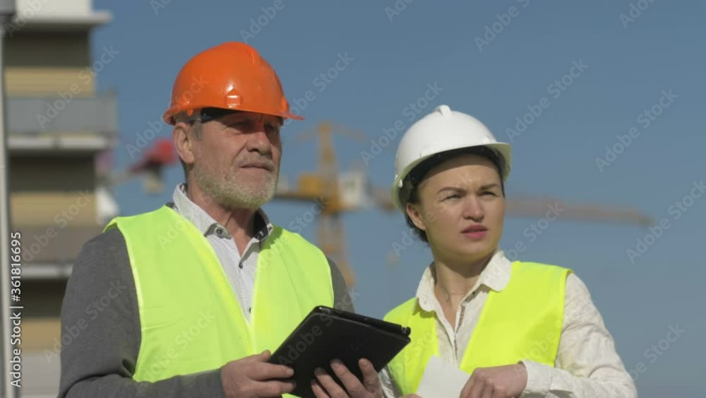 Elderly man with a tablet in his hands and a young woman at a construction site. Builders have fun discussing work issues. Elderly man with a tablet in his hands and a young woman at a construction site. Builders have fun discussing work issues.