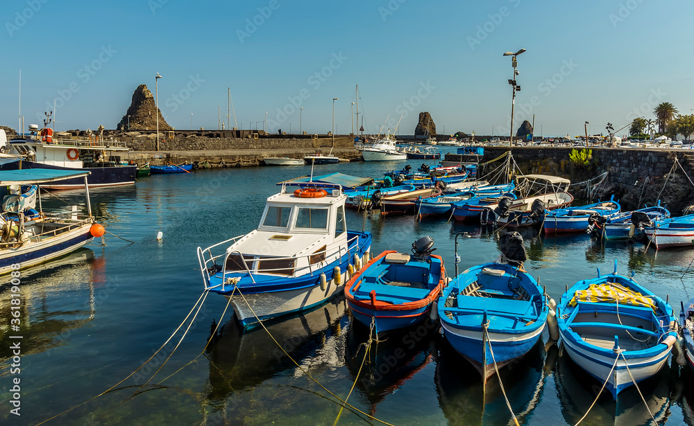 Fishing boats lined up in the harbour of Aci Trezza, Sicily with stacks of Isole dei Ciclopi in the background in summer