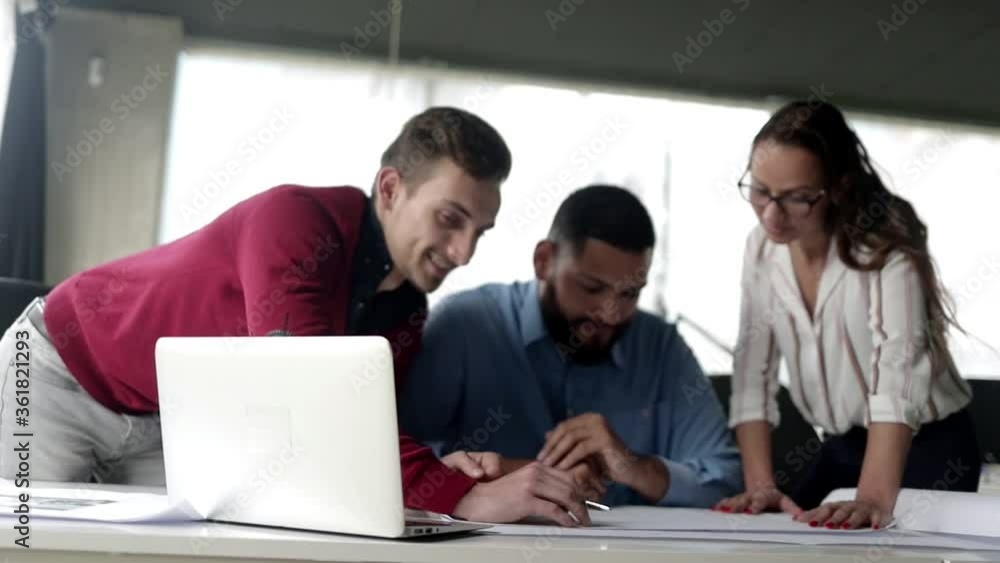 Team of experts discussing design project, working on blueprint at office workplace, looking at camera, posing and smiling. Low angle. Expertise or teamwork concept Team of experts discussing design project, working on blueprint at office workplace, looking at camera, posing and smiling. Low angle. Expertise or teamwork concept