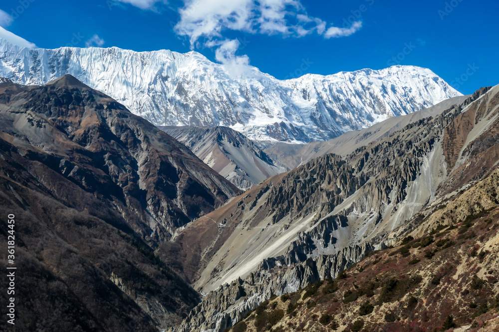 A panoramic view on a valley along Annapurna Circuit in Nepal. In the back there are high, snow capped Himalayan peaks. Slopes are overgrown with small bushes. Exploration and discovering