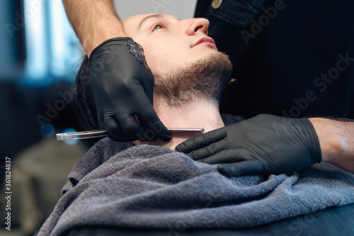 Professional barber shaves customer beard with straight razor. Beard cut with old-fashioned blade at barbershop. Handsome macho man getting his beard shaved in studio. Close-up shot.