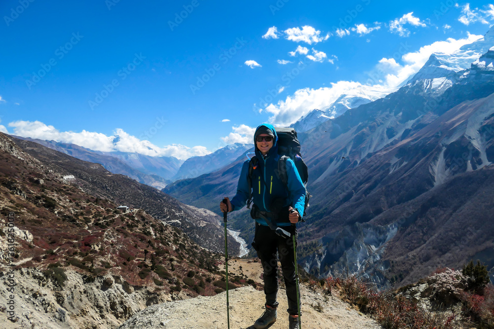 A man enjoying her trek to Tilicho Lake, along Annapurna Circuit in Nepal. In the back there are high, snow capped Himalayan peaks. Slopes are overgrown with small bushes. Exploration and discovering