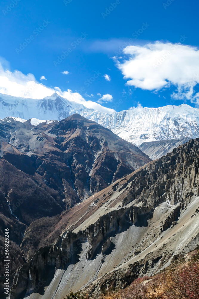 A panoramic view on a valley along Annapurna Circuit in Nepal. In the back there are high, snow capped Himalayan peaks. Slopes are overgrown with small bushes. Exploration and discovering
