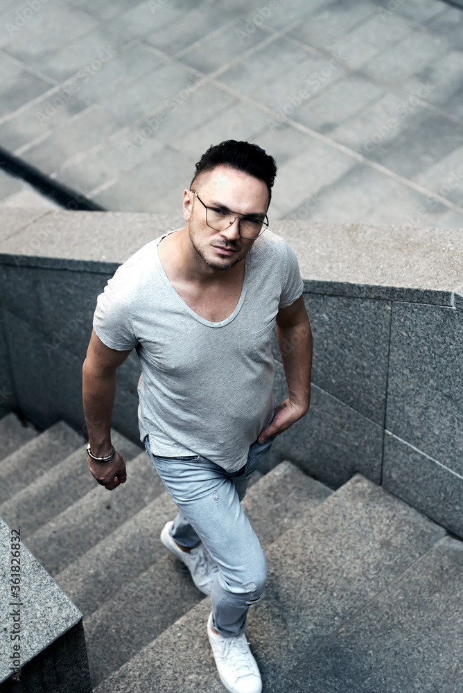 a stylish guy with glasses looks into the camera near and rises along the concrete steps to the top. strong strict male gaze. a fashionable gray T-shirt and light jeans. portrait in motion in the city
