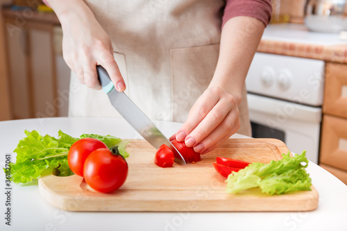 Female hands cut a juicy red tomato into slices with a knife on a wooden cutting board. A method of preparing vegetables and ingredients before cooking