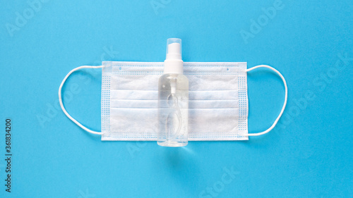 White medical mask and hand sanitizer in transparent bottle with spray cap at the middle of blue background. Simple flat lay with pastel paper texture. Medical concept. Stock photo.