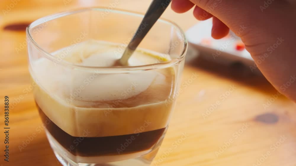 a glass with Vietnamese egg coffee is on a wooden table and a female hand is stirring a spoon of yellow foam from egg whipped yolk, slow motion. delicious sweet traditional north vietnamese drink.