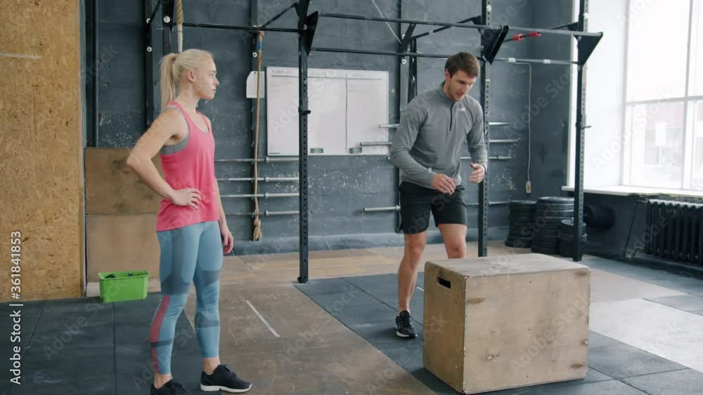 Young man is jumping on wooden box during crossfit training while female coach is watching sportsman. People, healthy active lifestyle and gym concept. Young man is jumping on wooden box during crossfit training while female coach is watching sportsman. People, healthy active lifestyle and gym concept.