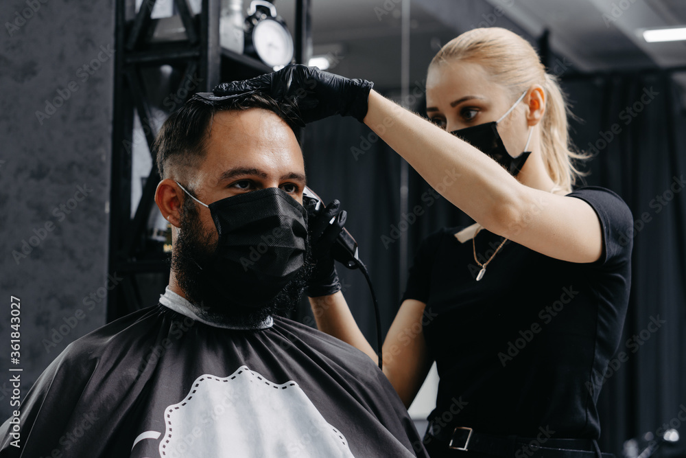 Woman barber cutting hair to a bearded man in face mask. Quarantine haircut concept.