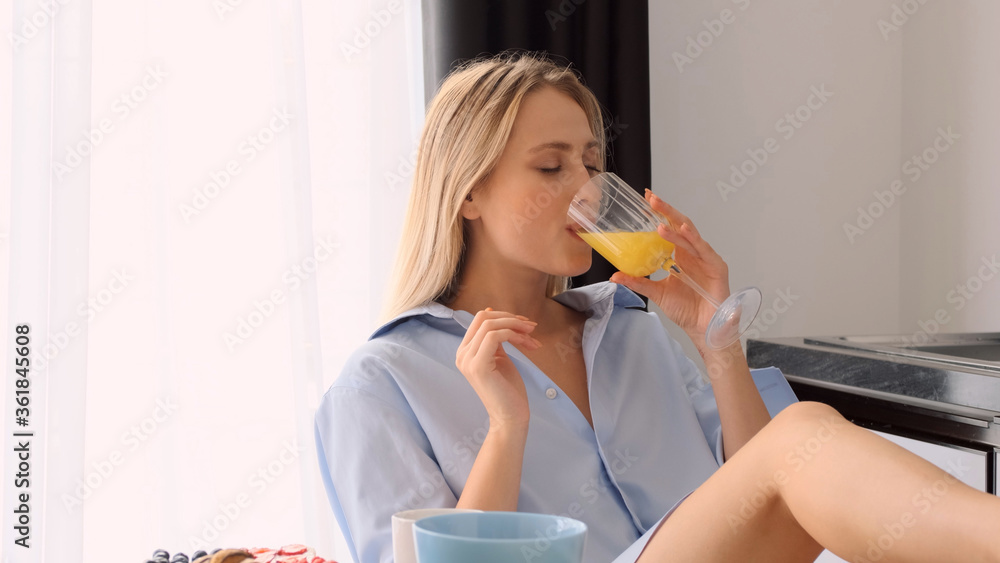 Beautiful blond girl in shirt drinking orange juice on kitchen. Attractive girl having breakfast at home Beautiful blond girl in shirt drinking orange juice on kitchen. Attractive girl having breakfast at home