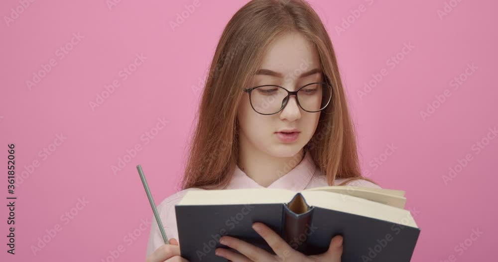 Concentrated girl in eyeglasses standing in studio with interesting book while new idea appeared in her head. Blonde female in pink shirt using pen for writing down all thoughts. Concentrated girl in eyeglasses standing in studio with interesting book while new idea appeared in her head. Blonde female in pink shirt using pen for writing down all thoughts.