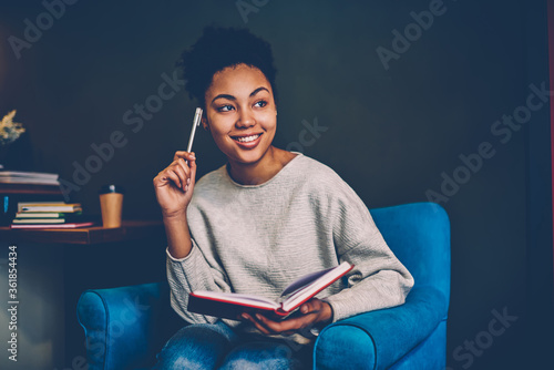 Εκτύπωση καμβά Smiling creative female blogger pondering on idea for publication sitting in comfortable armchair,young african american woman recalling information while planning to do list at home interior