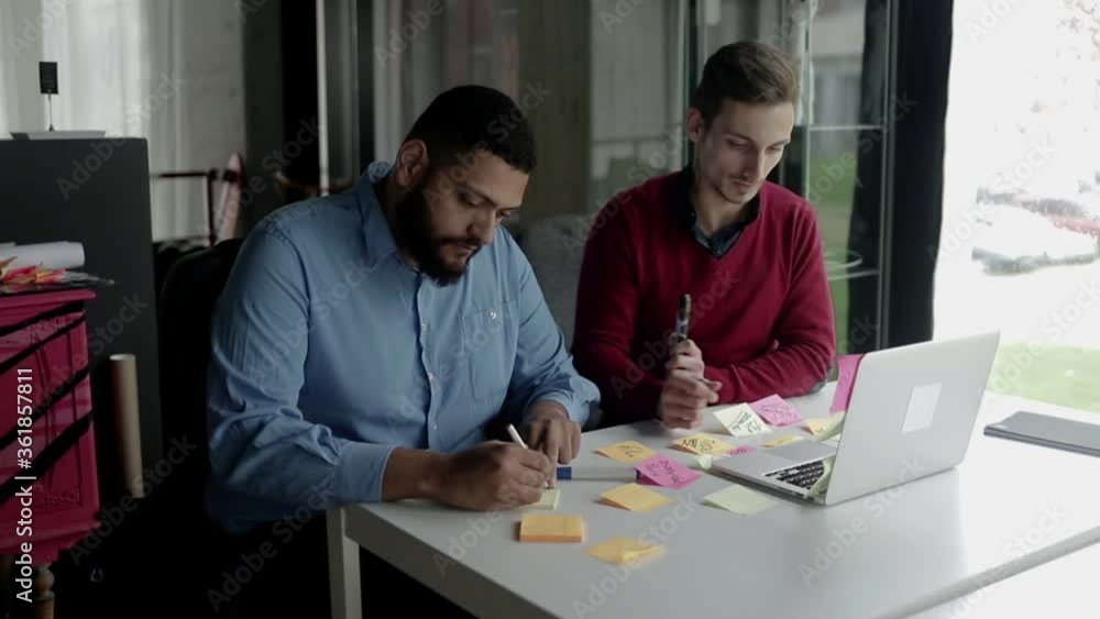 Diverse business men working on project, writing and sticking notes on desk, waving hello at laptop display. Medium shot. Business communication concept Diverse business men working on project, writing and sticking notes on desk, waving hello at laptop display. Medium shot. Business communication concept
