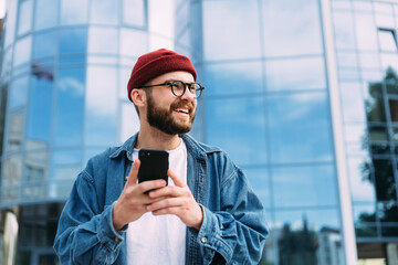 Portrait of happy handsome bearded young male hipster holding smartphone and looking to the side