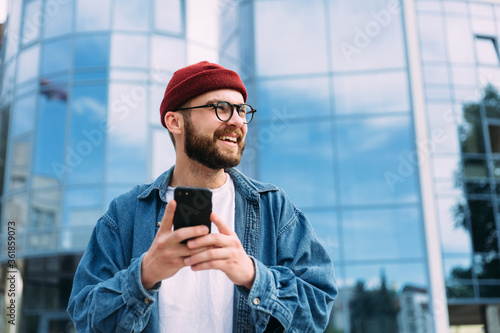 Portrait of happy handsome bearded young male hipster holding smartphone and looking to the side