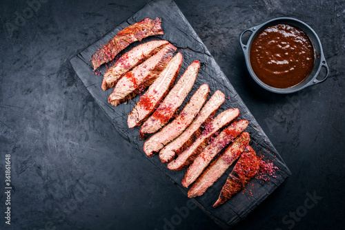 Traditional barbecue sliced dry aged wagyu flank steak offered with spicy BBQ Sauce and chili powder as overhead view on a rustic charred wooden cutting board with copy space left