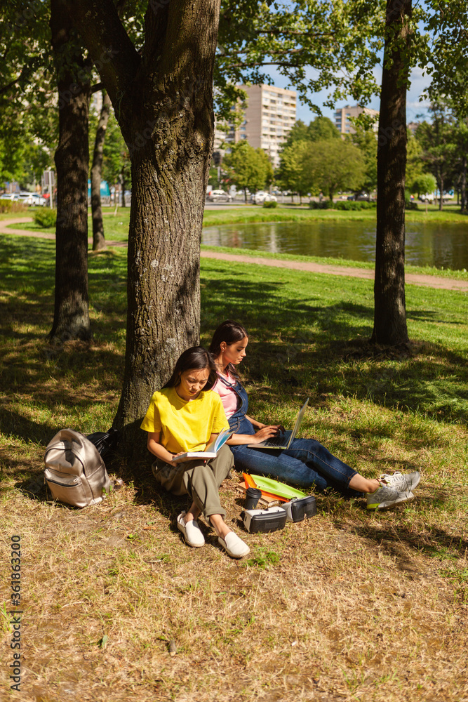 Two female college students sitting in shade of tree leaning on trunk in park. Asian girl reading textbook, her Caucasian friend using laptop. Backpacks, binders and vr headsets near them Two female college students sitting in shade of tree leaning on trunk in park. Asian girl reading textbook, her Caucasian friend using laptop. Backpacks, binders and vr headsets near them