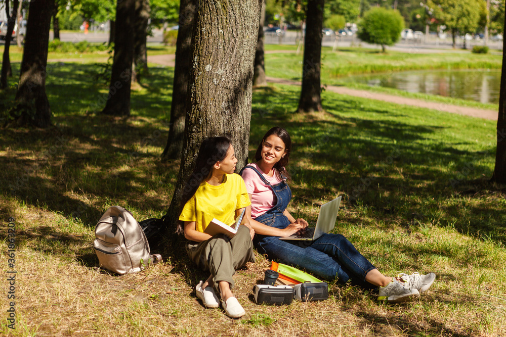 Two female college students sitting in shade of tree in park and talking looking at each other. Asian girl reading book, her Caucasian friend using laptop. Backpacks and vr headsets near them