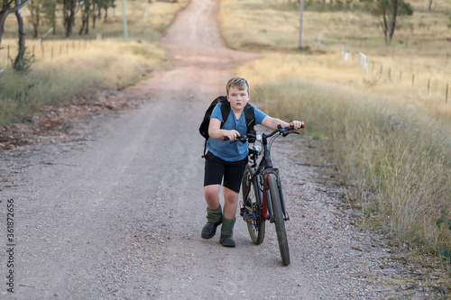 Young boy pushing bike up hill on gravel road in dry countryside