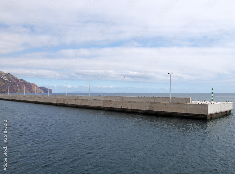 the long concrete harbour wall surrounding the marina and docks in funchal madeira with the buildings of the town in the distance
