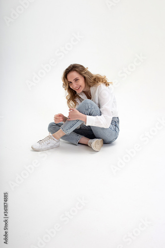 portrait of young smiling caucasian woman posing in shirt, blue jeans, sitting on white studio background holding knees. model tests of pretty girl in basic clothes. attractive female poses, laughs