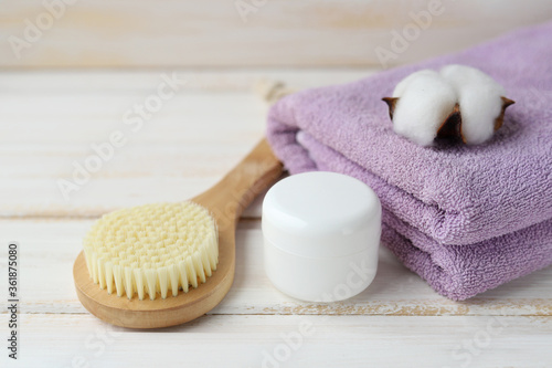 Spa treatments in the sauna. A wooden massage brush for cellulite prevention, a white jar with skin care product, a purple Terry towel and a cotton flower on a white wooden background. Selective focus