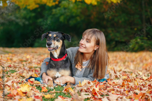 Young Caucasian woman lying on ground fall leaves hugging dog. Owner walking with pet on autumn day. Best friends having fun outdoors. Friendship of human with domestic animal. © anoushkatoronto