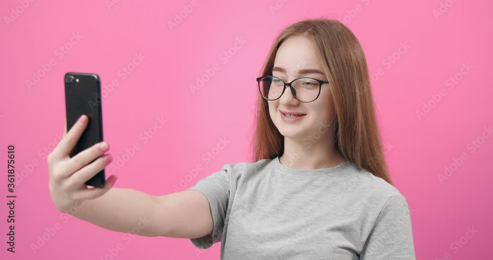 Smiling pretty woman in eyeglasses having video chat on smartphone while posing in studio with pink background. Young lady with blond hair talking with friends through video call. Smiling pretty woman in eyeglasses having video chat on smartphone while posing in studio with pink background. Young lady with blond hair talking with friends through video call.