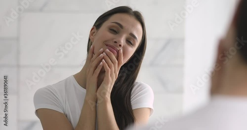Happy confident attractive young woman touch pretty face after applying cream do make up look in mirror enjoy healthy clean soft moisturized hydrated skin care admire herself feel proud narcissistic