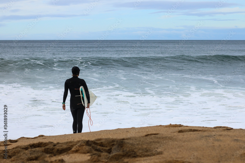 an unidentifiable woman in a wetsuit holiding her surfboard waiting for the right time to join the surf, coastal Victoria, great ocean road, Australia an unidentifiable woman in a wetsuit holiding her surfboard waiting for the right time to join the surf, coastal Victoria, great ocean road, Australia