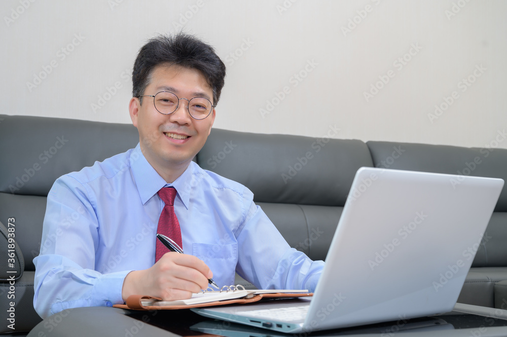 Middle-aged Asian businessman sitting on sofa in living room working at home. Telecommuting concept.