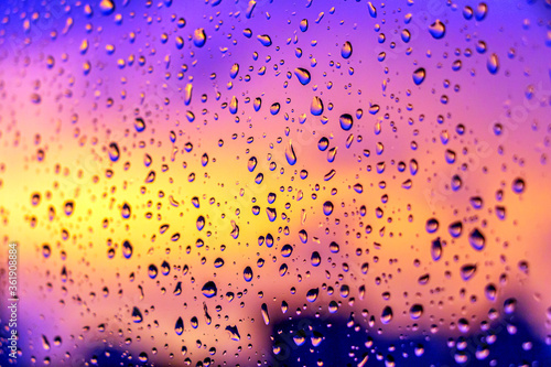 Bright fiery sunset through raindrops on window with bokeh lights. Abstract background. Water drop on the glass against the blurred silhouettes high-rise city.