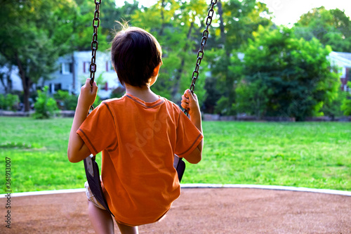 Boy, child sitting on a swing in a playground and looking off into the distance. View from behind, rear.