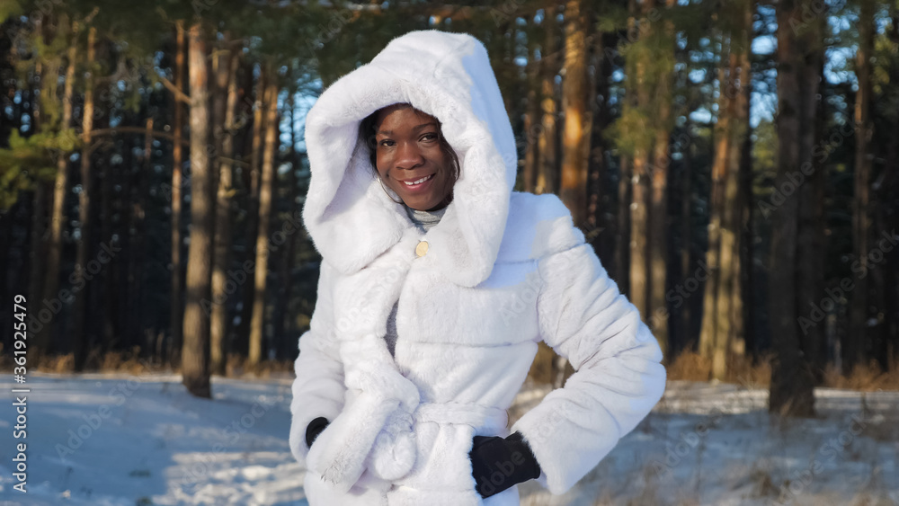 Obraz premium smiling African-American girl in white fur coat dresses hood in winter park closeup