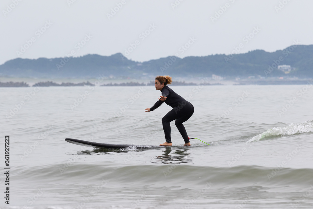 Young multiracial African American lady with amazing smile, freckles & frizzy hair, learning to surf wearing a black wetsuit on a black surfboard. She is Surfing in Chiba Japan near to Ichinomiya.