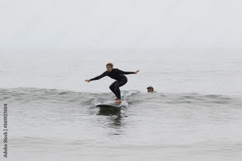 Young multiracial African American lady with amazing smile, freckles & frizzy hair & an Asian Japanese Surf Instructor having a surf lesson together in Chiba, Japan They are wearing black wetsuits.
