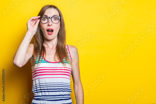 Surprised young girl holding glasses in a striped dress on a yellow background