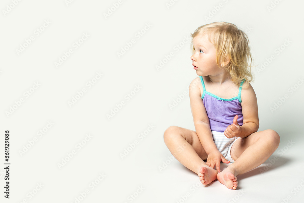 Beautiful cute little baby sits and looks away and makes a class gesture in a T-shirt and panties on a white background Beautiful cute little baby sits and looks away and makes a class gesture in a T-shirt and panties on a white background
