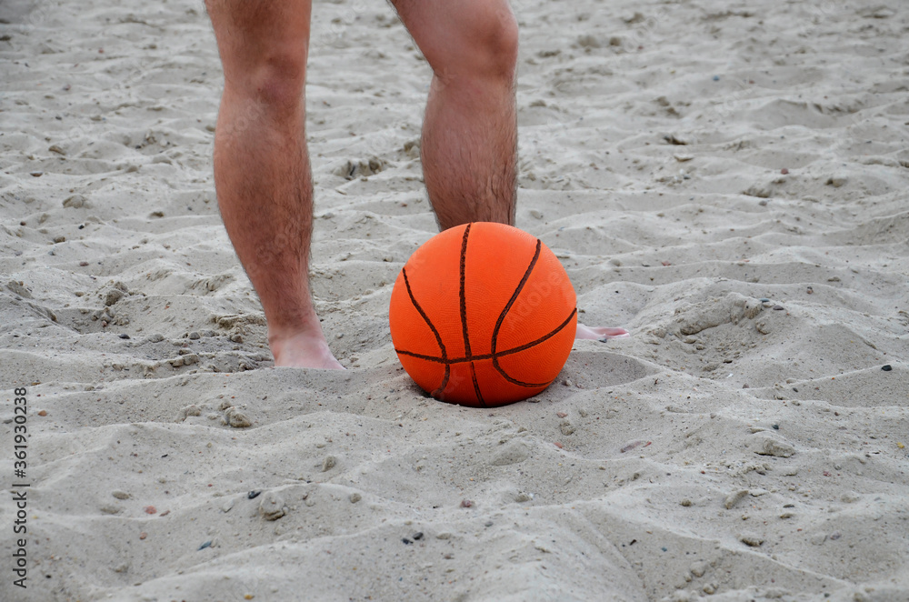 A basketball is lying on the sand against the background of men's feet. A young man with a ball on the beach. Bottom view. Summer period. Rest.
