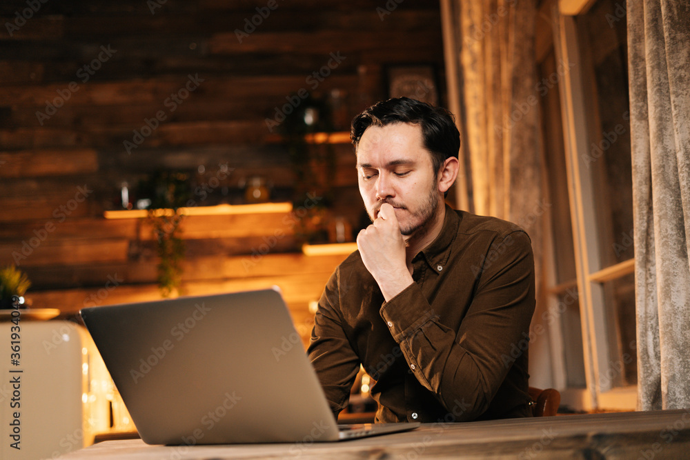 Thoughtful young man in casual clothing working on laptop while sitting at the table in cozy living room. Concept of remote working from home. Thoughtful young man in casual clothing working on laptop while sitting at the table in cozy living room. Concept of remote working from home.