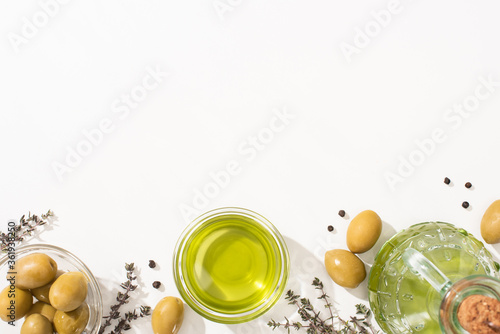 top view of olive oil in bowl and bottle near green olives, herb and black pepper on white background