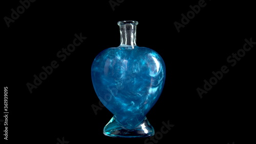 Glass bottle in the form of a heart with blue liquid. The elixir is spinning and overflowing with liquid. Potion of love is isolated on a black background.