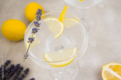 Infused water with ice and lemon in a glass stands on a gray concrete background. The glass is decorated with a sprig of lavender and a slice of lemon. Nearby are two lemons. Close-up, top view