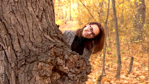 A young woman in glasses hides behind a large tree of leaves in the autumn forest. Yellow leaves around
