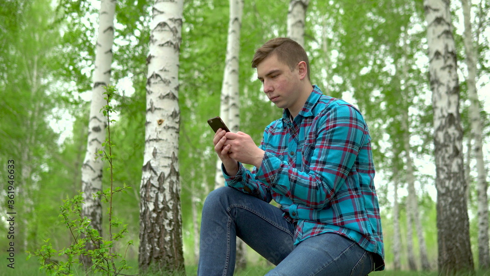 Obraz premium Young man in nature with a phone in his hands. A man sits on a stump in a birch forest and leads works through a smartphone.