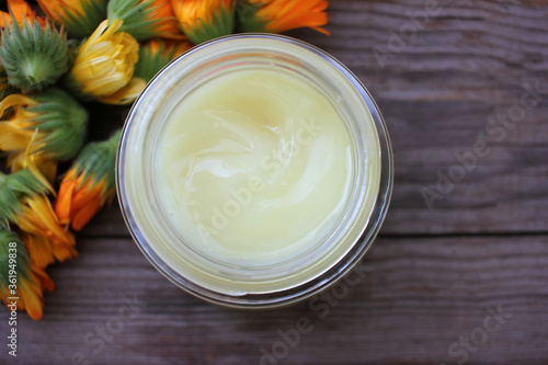Calendula flowers and diy face cream in glass jar on wooden table background with copy space. Homemade natural organic cosmetics with medicinal herbs.