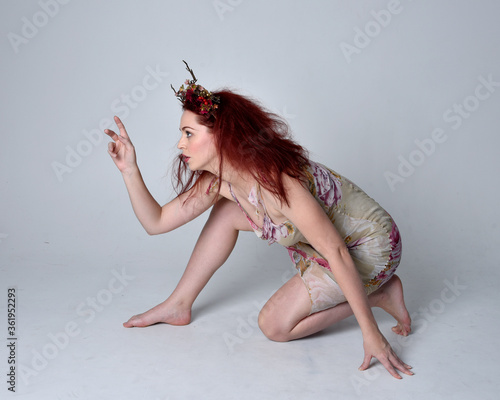full length pose of red hair girl wearing floral fairy dress and headdress. sitting pose on grey studio background.